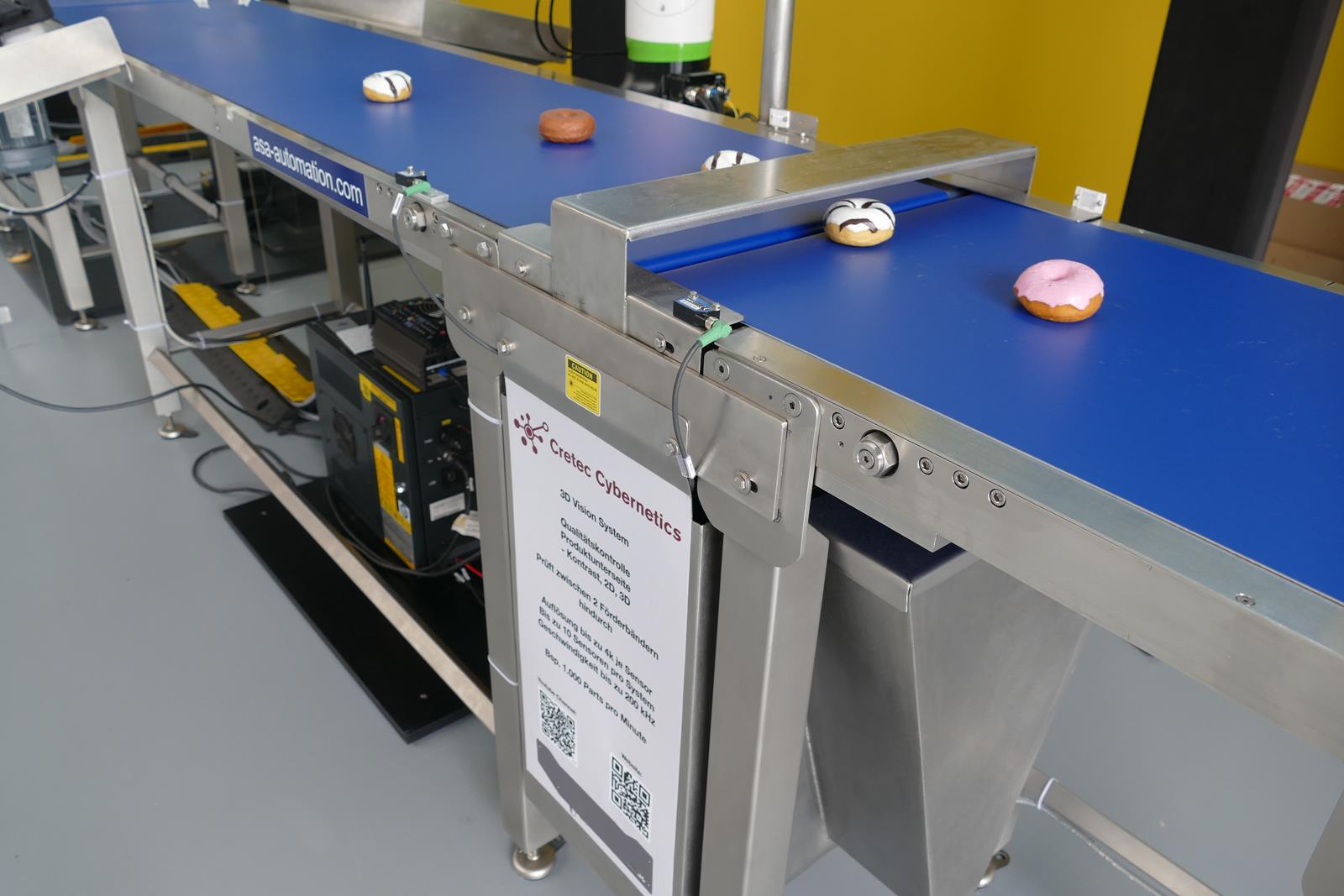 An der ersten Bildverarbeitungsstation reicht ein ganz schmaler Spalt zwischen zwei Transportbändern aus, um die Donuts mit einem 3D-Profilsensor der Z-Trak2-Serie von Teledyne Dalsa auf Fehler an der Unterseite zu überprüfen. Bildquelle: Teledyne DALSA An der ersten Bildverarbeitungsstation reicht ein ganz schmaler Spalt zwischen zwei Transportbändern